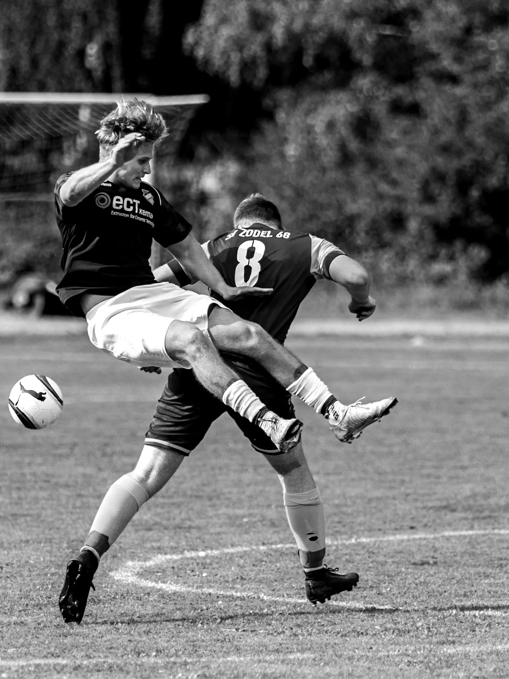 Schwarzweiß: Ein Spieler springt im Zweikampf über das Bein seines Gegenspielers.