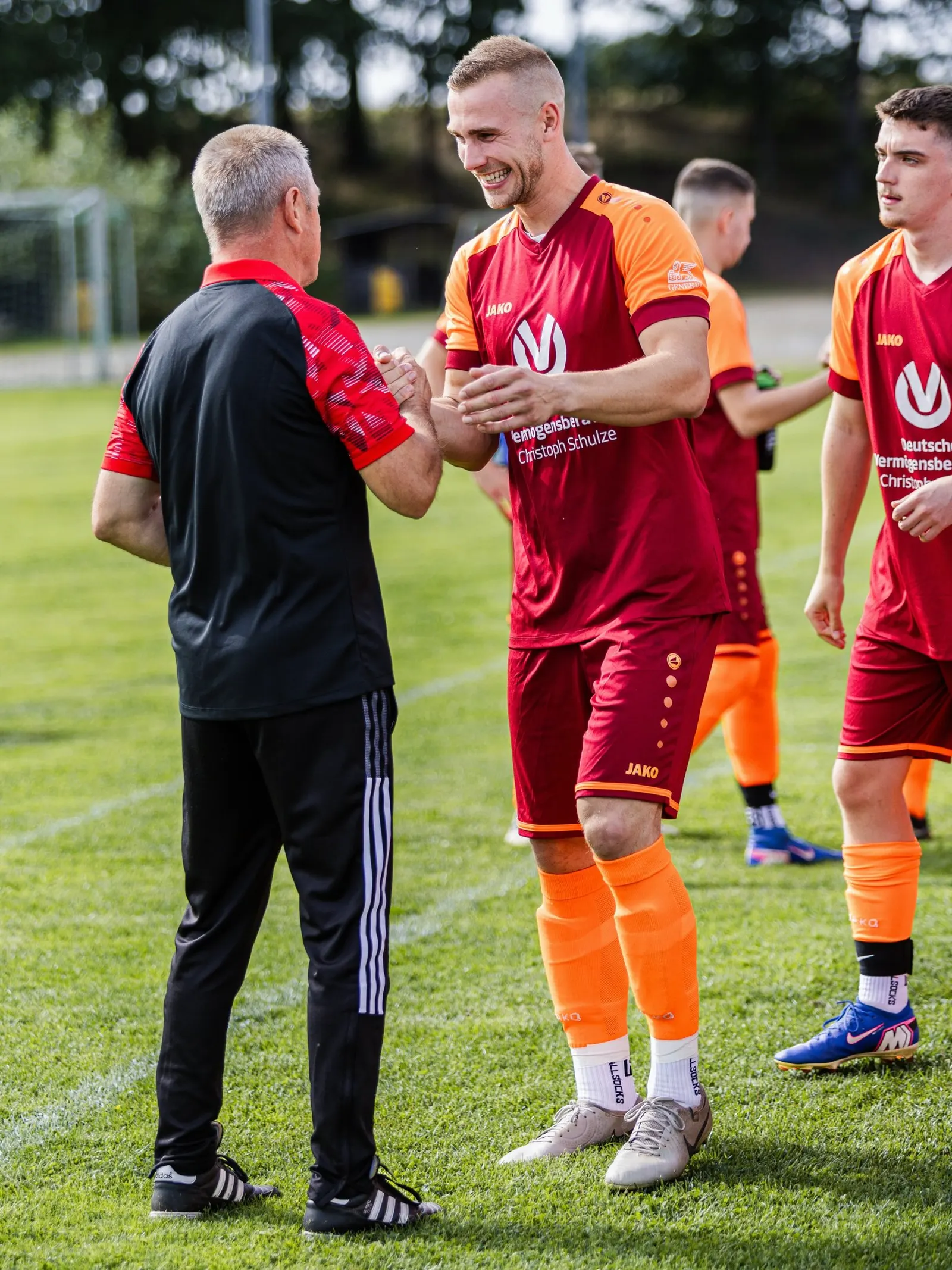 Der Trainer spricht lachend mit zwei Spielern am Spielfeldrand.