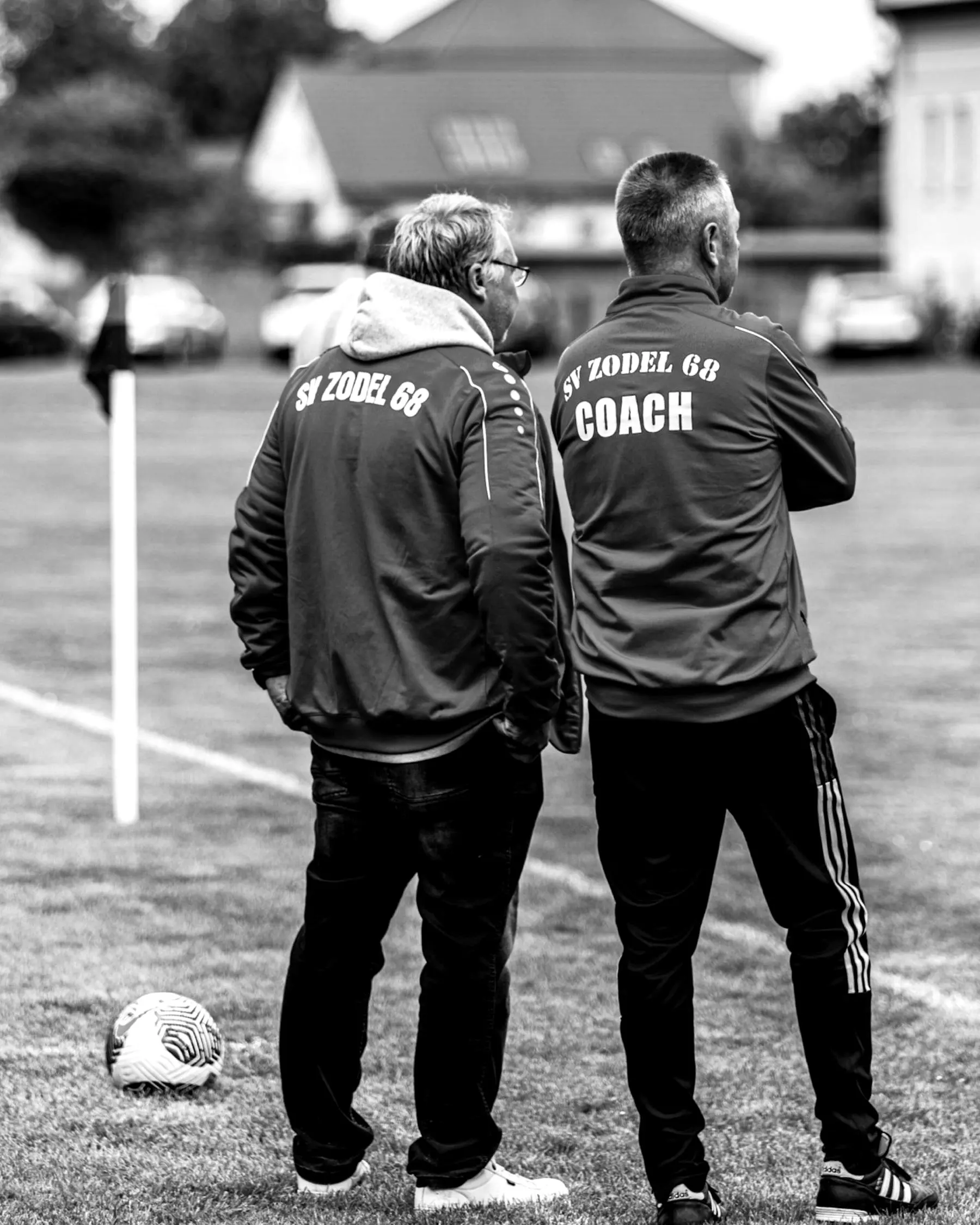 Zwei Trainer in roten Jacken beraten sich am Spielfeldrand.